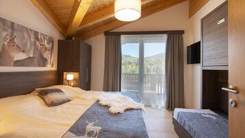 Ein Schlafzimmer mit einem großen Bett, einem kleineren Bett, einer Holzbalkendecke, einem Hirschgemälde, einem Fernseher und einem Balkon mit Vorhängen.