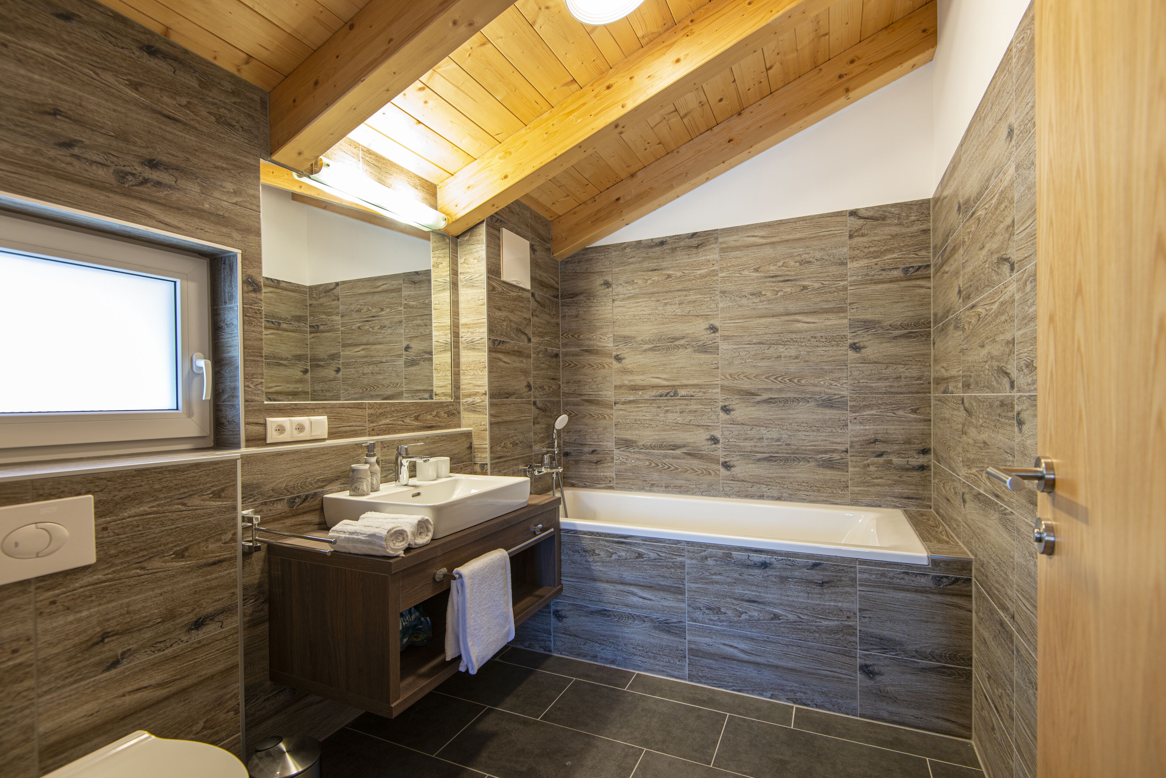 A modern bathroom with a wooden ceiling, a large mirror, a sink, a bathtub, and dark wood-patterned walls.