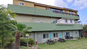 Un edificio de dos plantas con fachada verde, zona de estar al aire libre y un pequeño jardín con palmeras.