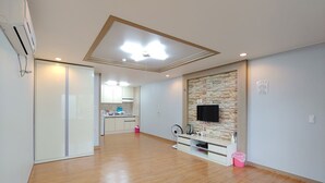 Una habitación amplia con piso de madera, una kitchenette con armarios blancos, un televisor integrado y una lámpara en el techo.