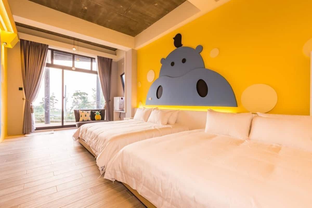 A hotel room with two beds, a large window with curtains, a wall mural, and a wooden floor.