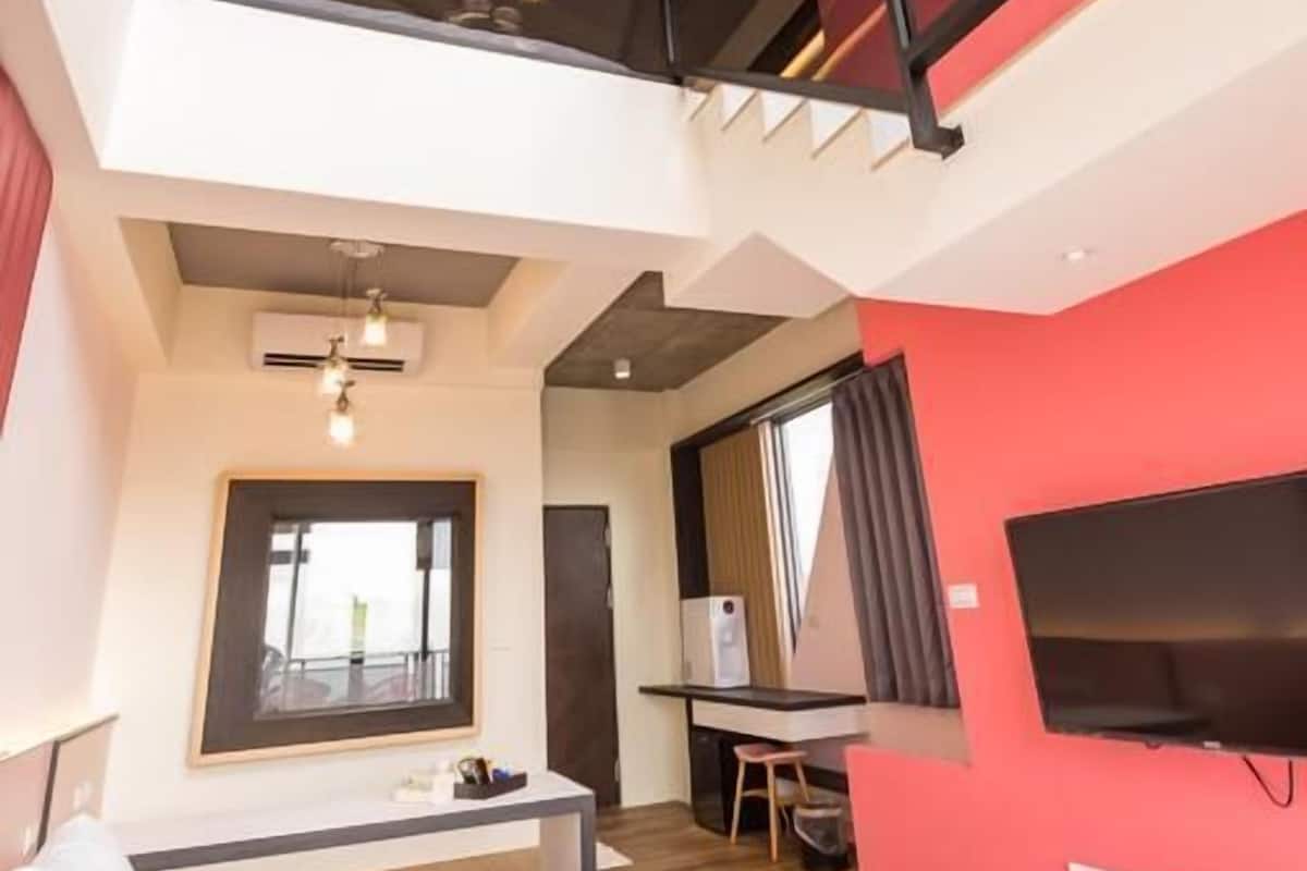 A modern hotel room with a bed, desk, and a large TV. There is a balcony with a railing and a staircase.