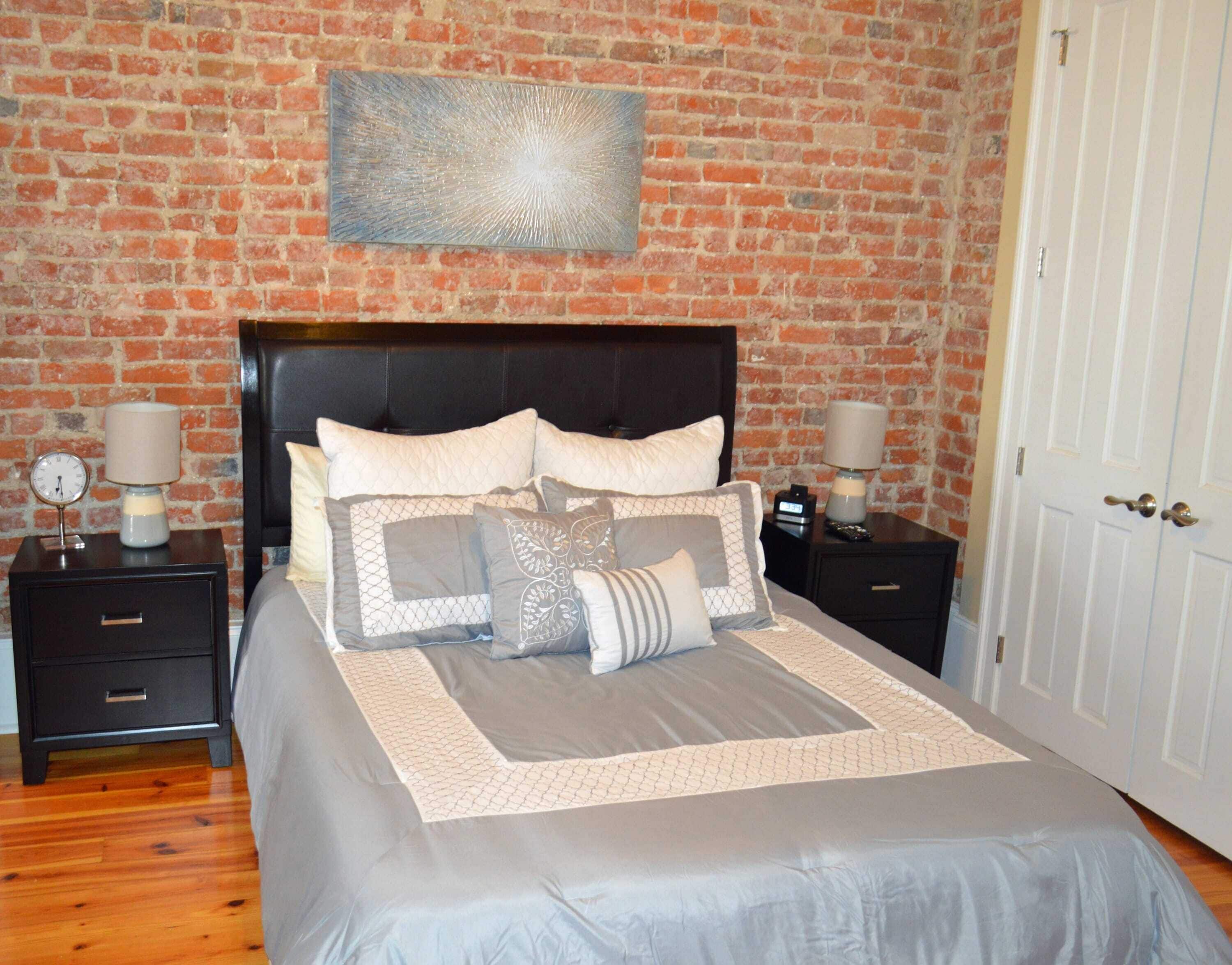 Un dormitorio con pared de ladrillo, cabecera negra, cama con varios almohadones, dos mesitas de noche con lámparas y una puerta blanca.