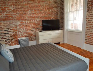 A room with a brick wall, a flat-screen TV on a white dresser, a bed with a grey mattress, and a window with blinds. - "Margaux" Fully renovated 2 bed / 2 bath  Walk to WWII, Superdome, Convention Center, Cruise Port (New Orleans)