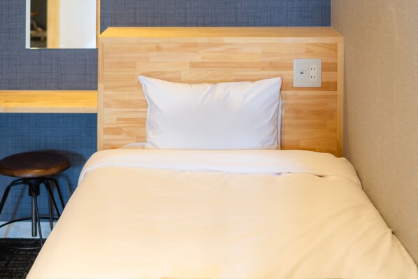A neatly made bed with a wooden headboard and a white pillow, a small stool, and a power outlet on the wall.