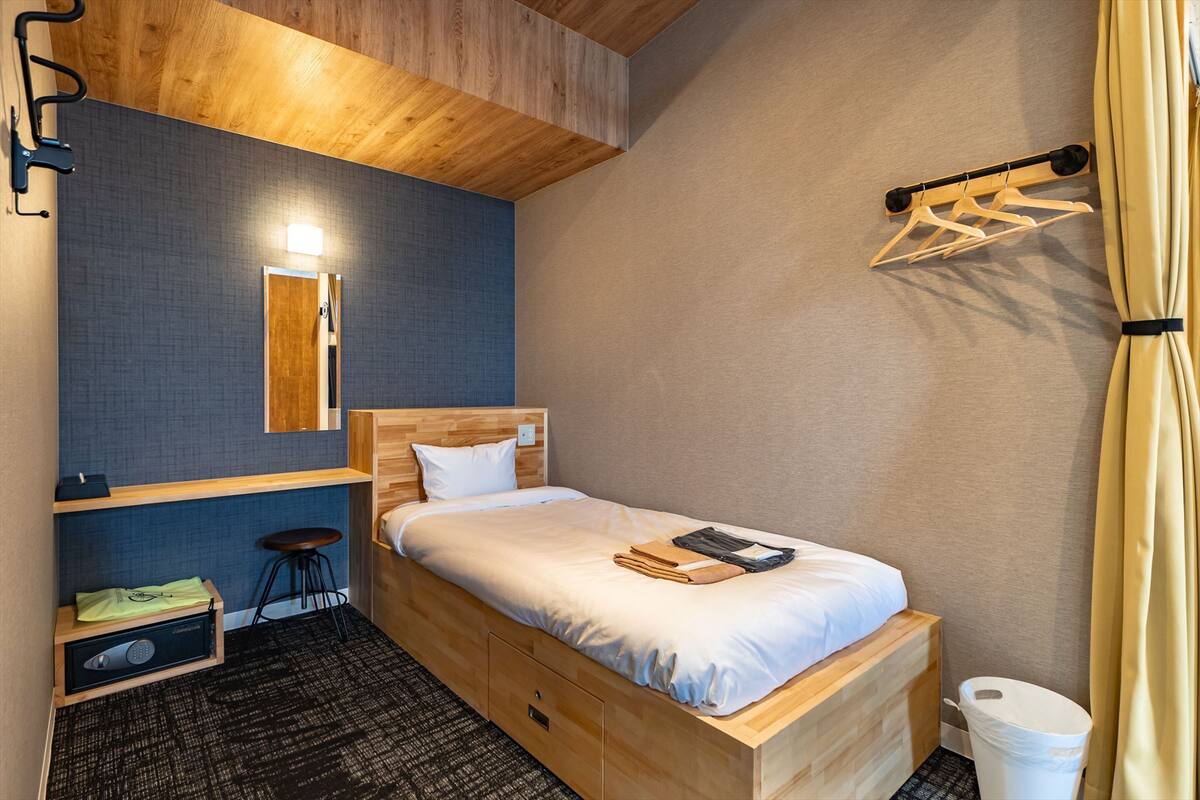 Una habitación de hotel pequeña con una cama individual, un taburete, una mesita de noche de madera, un espejo y una lámpara colgante.