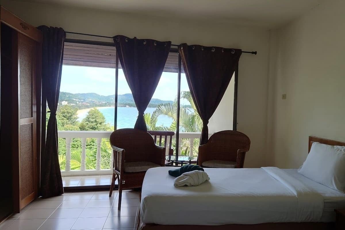 A hotel room with a bed, two chairs, a nightstand, a lamp, and a view of the sea and mountains.