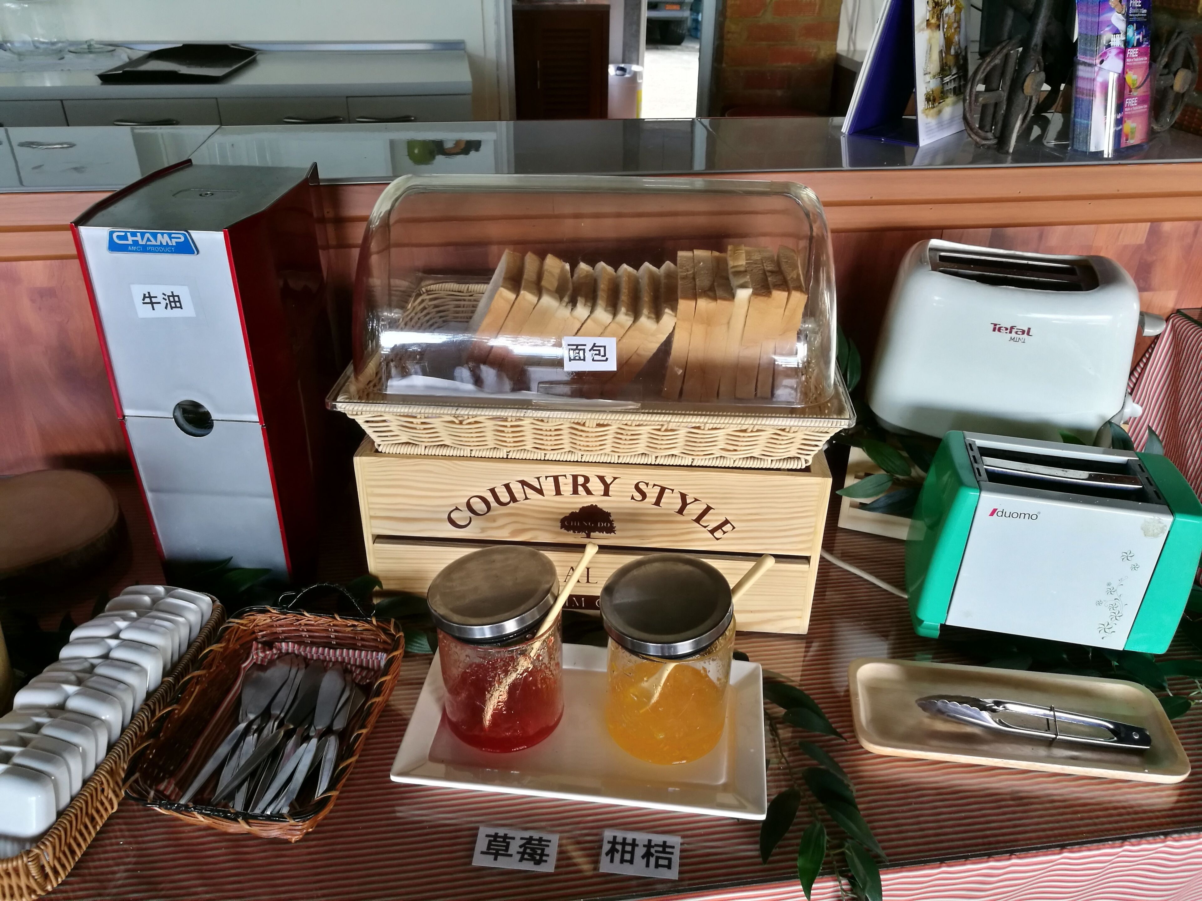 A buffet setup with a variety of food items, including a wooden box labeled "COUNTRY STYLE," a toaster, and two jars of jam.