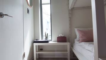 Um quarto pequeno e bem iluminado com uma cama beliche, uma mesa de cabeceira com um estojo de lenços e uma pequena planta em vaso, e uma janela que oferece luz natural.