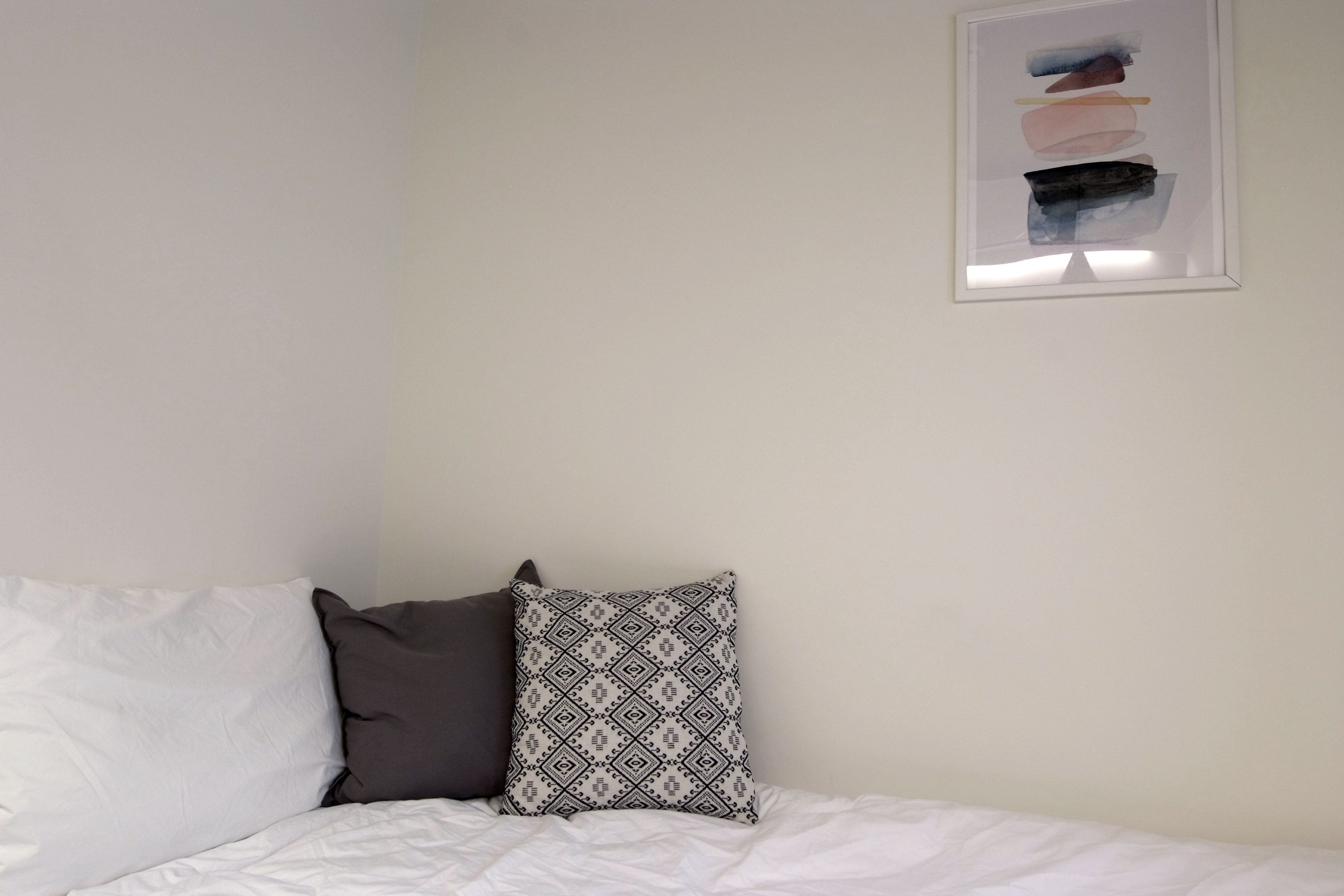 A bed with white bedding and two pillows, one patterned and one solid dark color, against a plain wall with a framed abstract artwork above.