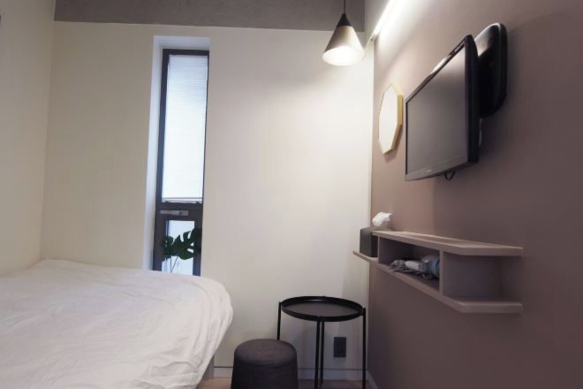 A modern hotel room with a bed, a small round table, a wall-mounted TV, and a window with a view.