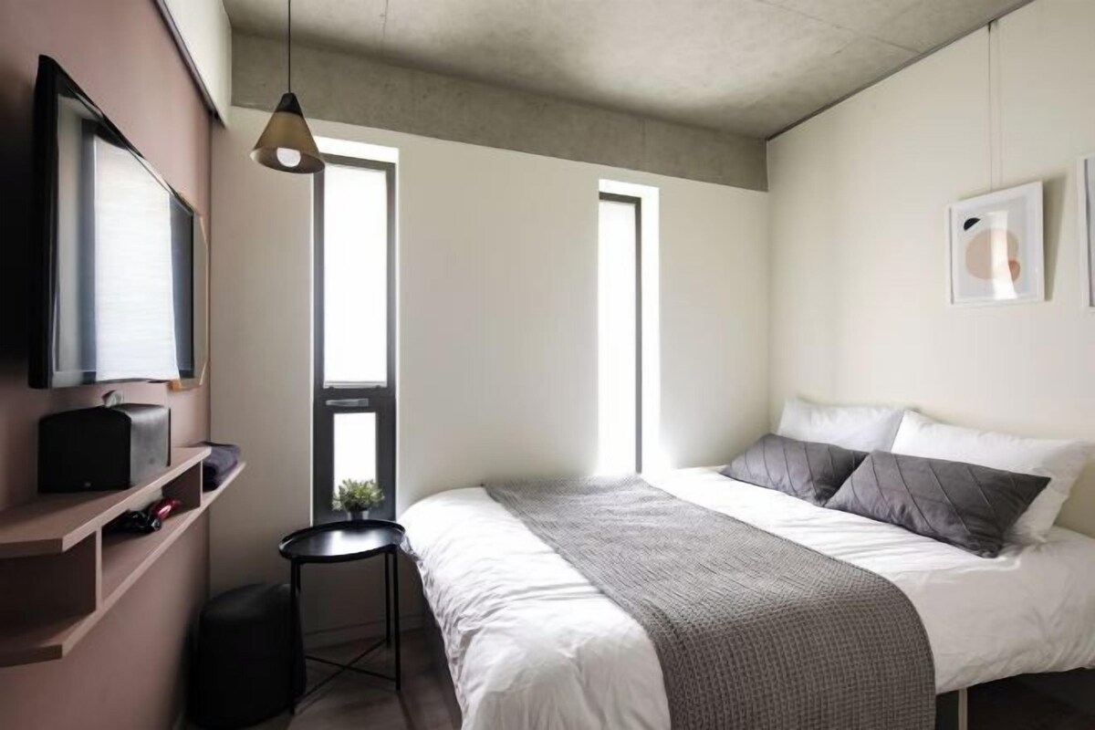 A modern bedroom with a bed, a TV mounted on the wall, and a window providing natural light.