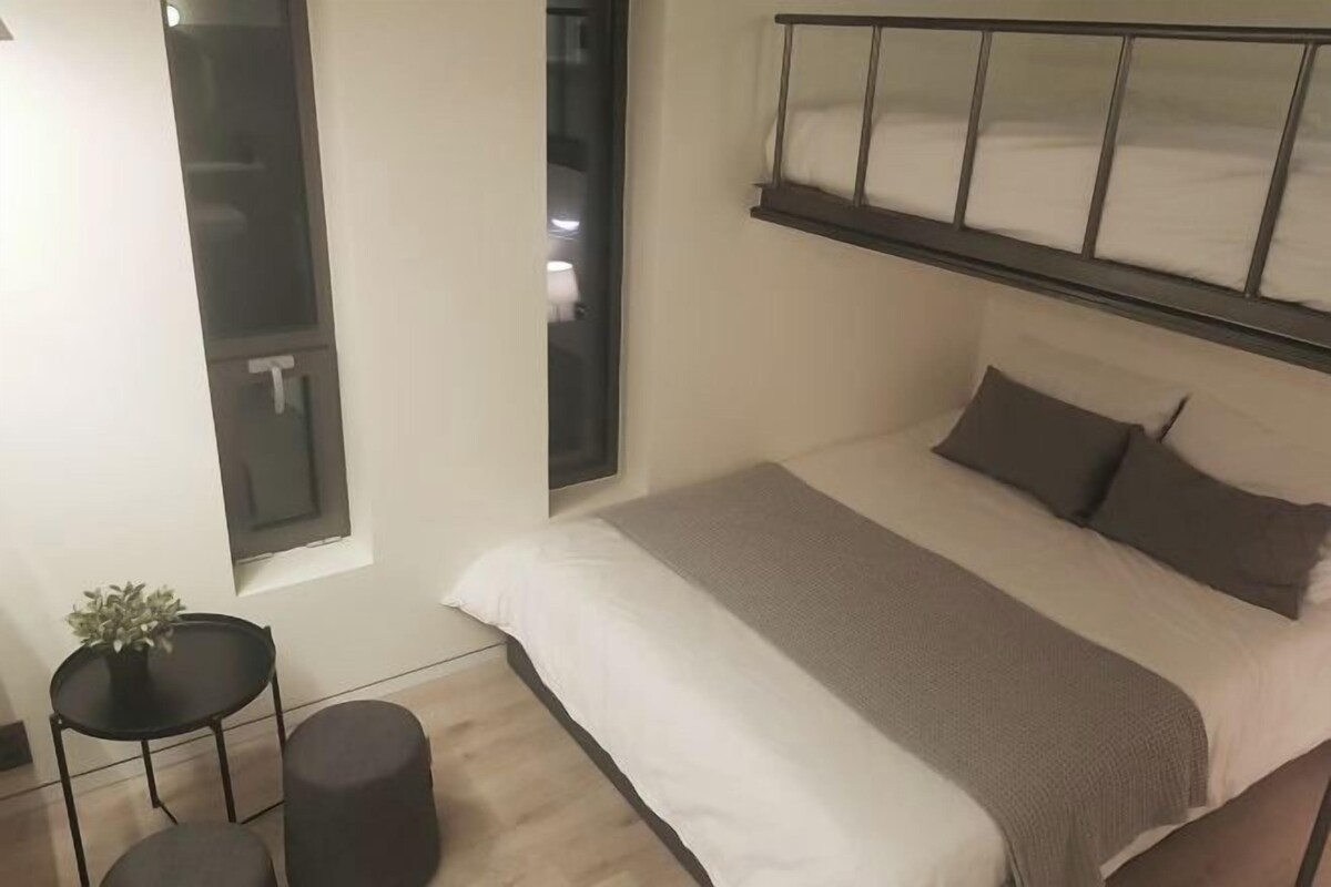 A bedroom with a bed, a bedside table, a small stool, and a window.