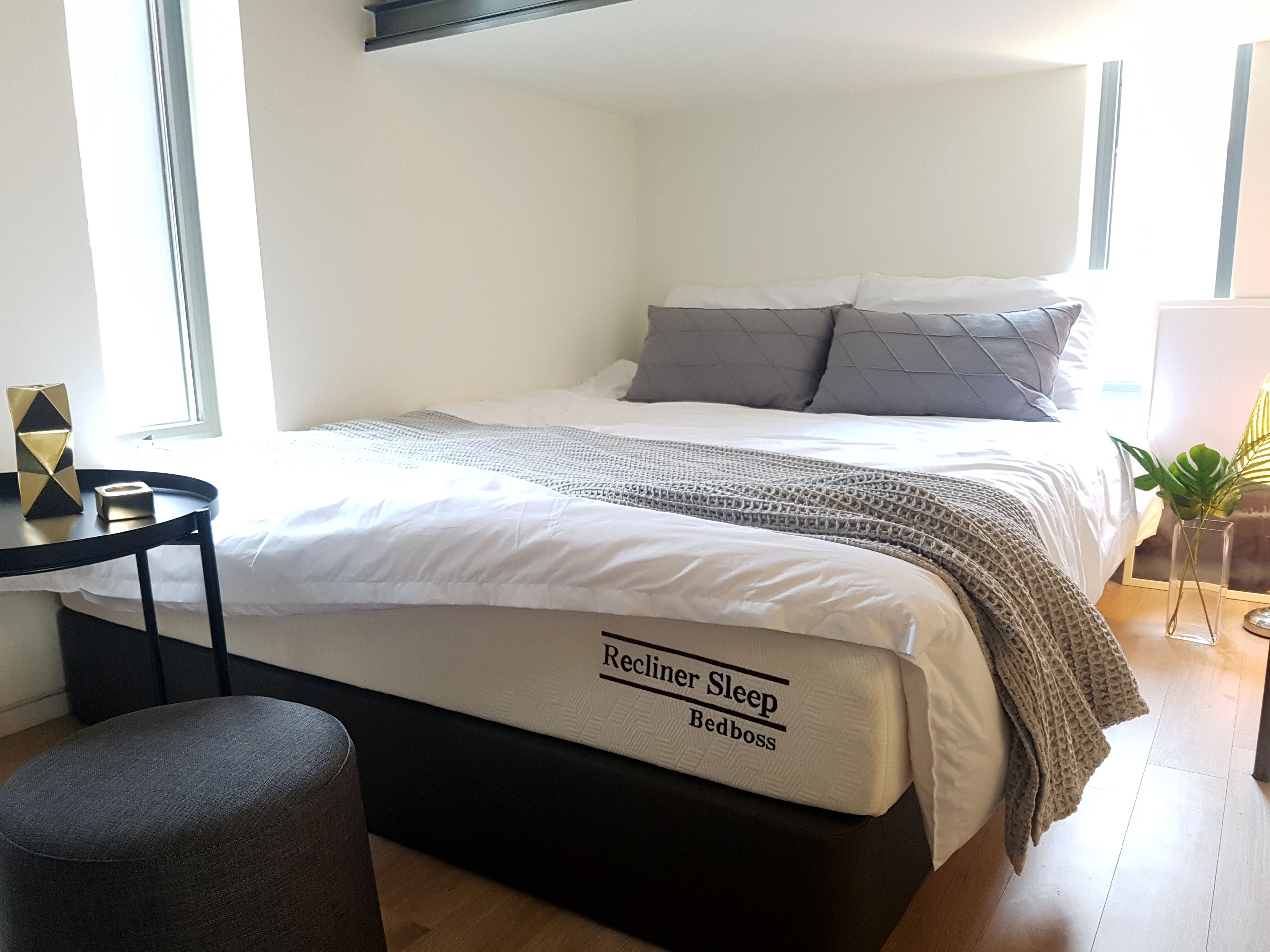 A neatly made bed with a branded mattress, a bedside table with a lamp, and a small potted plant.