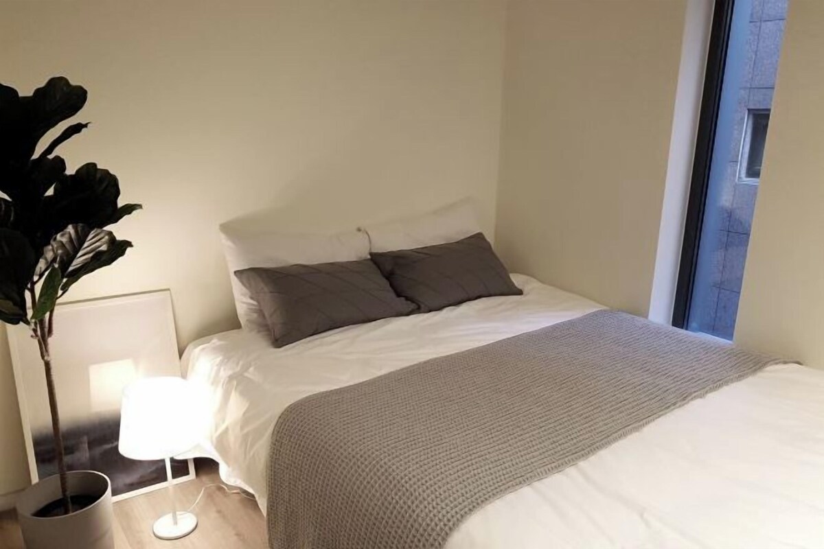 A neatly made bed with a gray blanket, a white pillow, and a dark gray pillow. There is a potted plant to the left of the bed and a white lamp on the floor beside it.