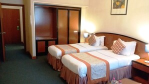 Habitación de hotel con dos camas, un armario, una mesita de noche con lámpara y una pequeña mesa con botellas y un control remoto.