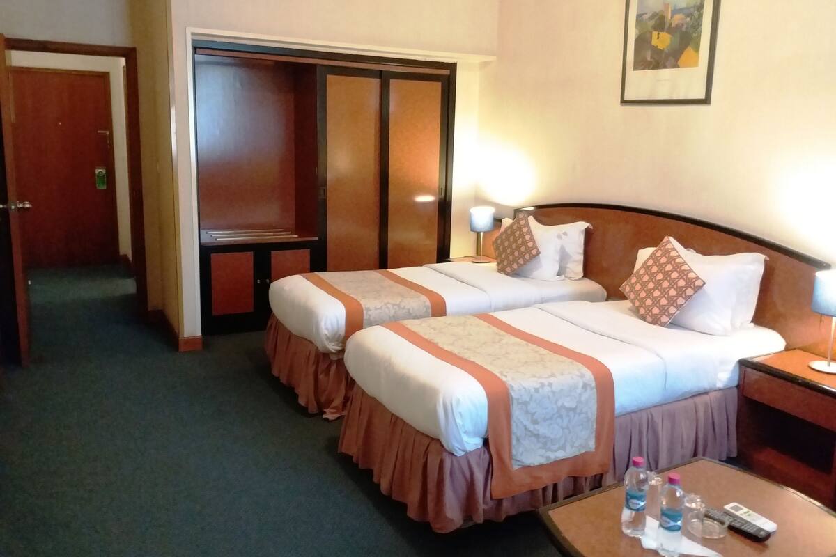 Une chambre d’hôtel avec deux lits, une armoire, une table de chevet avec une lampe et une petite table sur laquelle se trouvent des bouteilles et une télécommande.