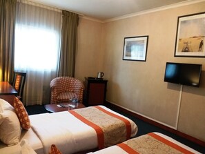 Habitación de hotel con dos camas, una silla, una mesita, un televisor y una ventana con cortinas.