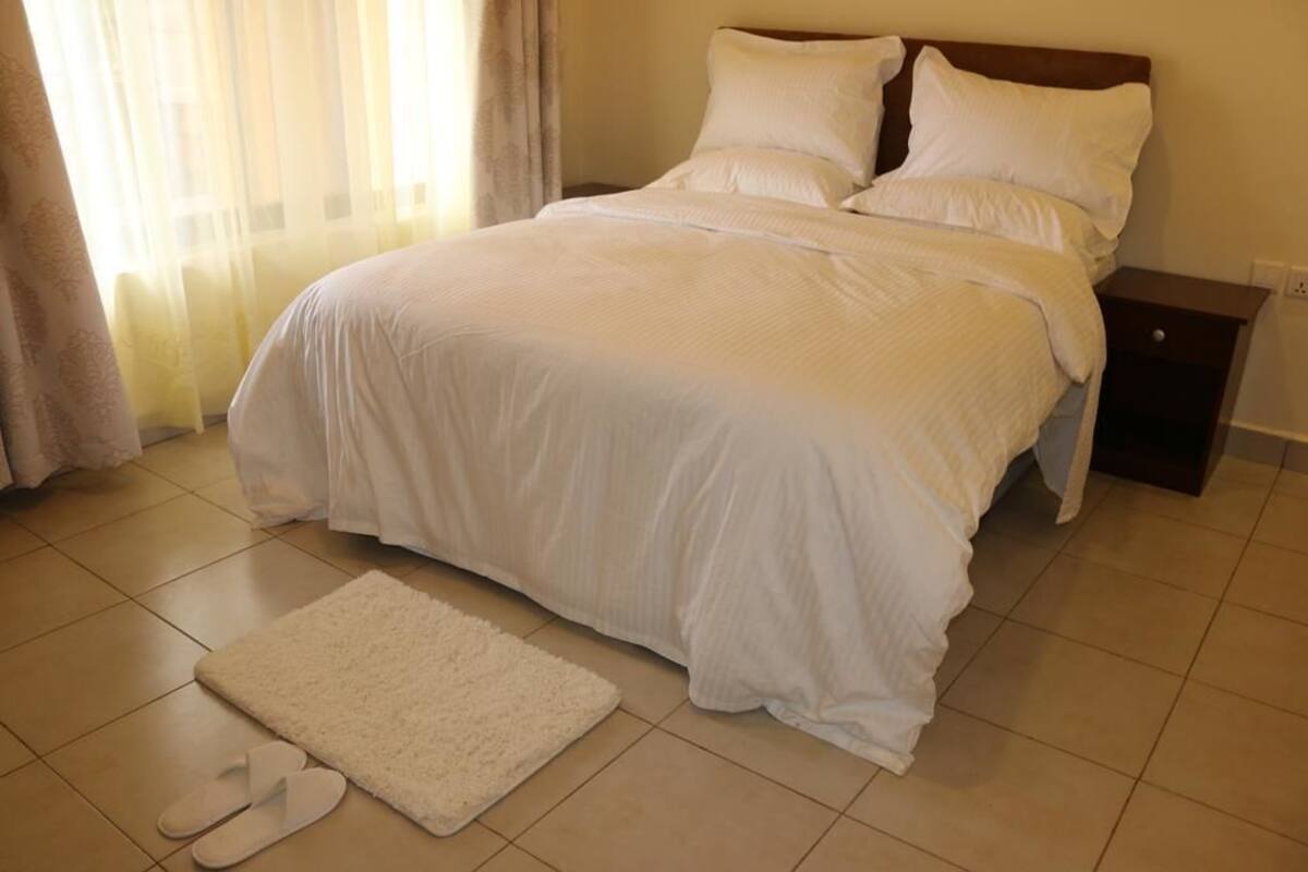 A neatly made bed with white bedding, a white rug, and a pair of slippers on the floor.