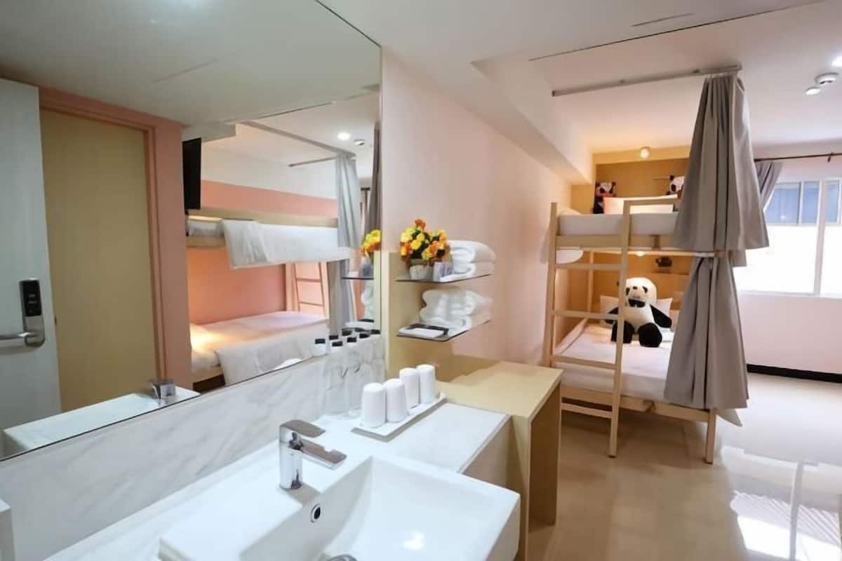 A hotel room with a bunk bed, a bathroom with a large mirror and two sinks, and a bedside table with toiletries.