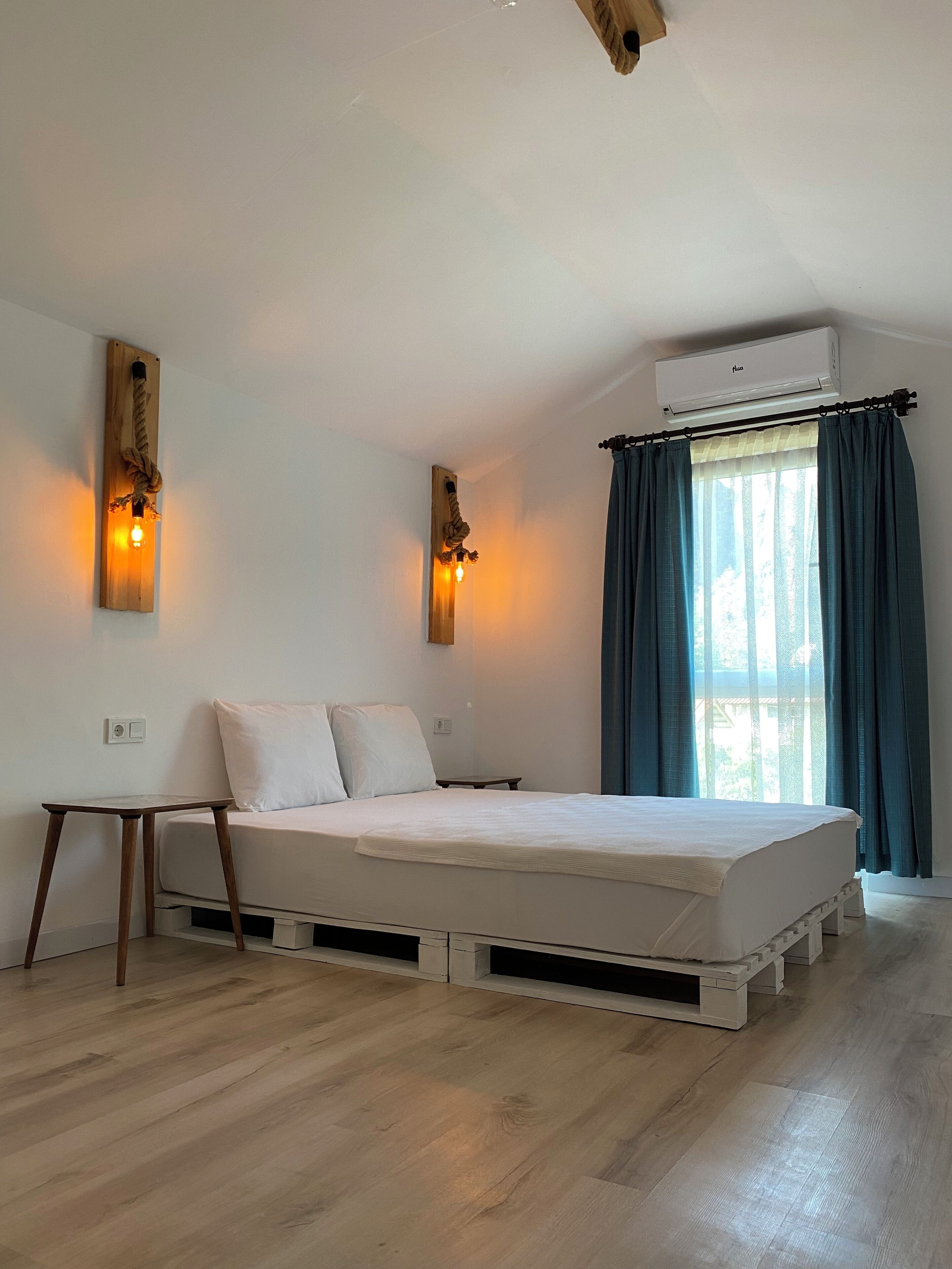 A bedroom with a bed, wooden floor, wall-mounted air conditioning unit, and two wall sconces.