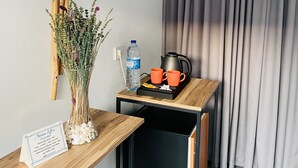 Una habitación compacta con un escritorio de madera, una mesita con una tetera, tazas y una botella de agua. Un jarrón con flores secas está sobre el escritorio, y se observa un cartel con información de contacto.
