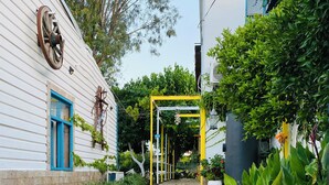Un sendero pavimentado conduce a una puerta amarilla, bordeado por abundante vegetación y un edificio blanco con un ventanal azul.