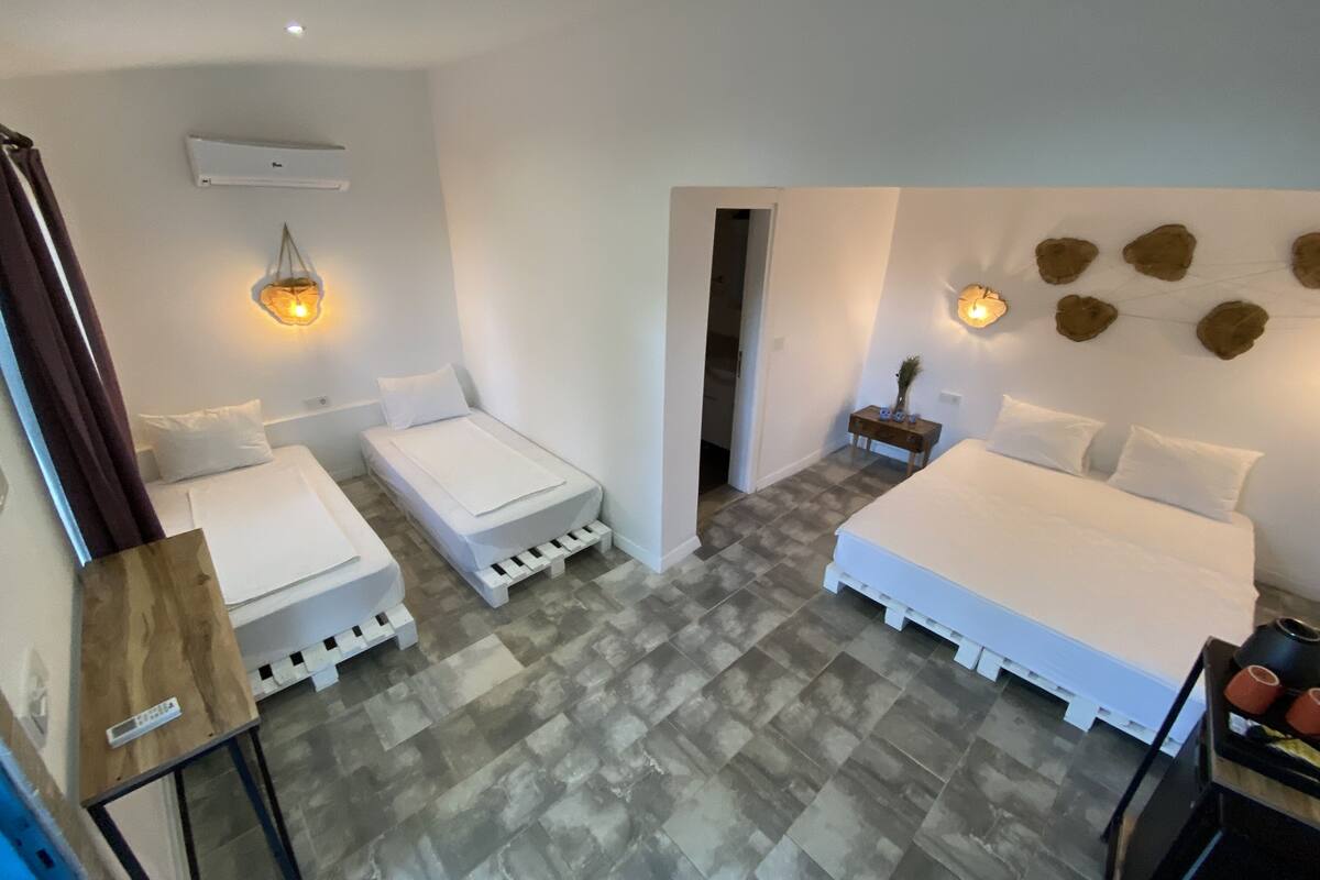 Un dormitorio con dos camas individuales, una cama doble más grande, una mesita de noche de madera, un aplique de pared y una pequeña mesa con un control remoto.