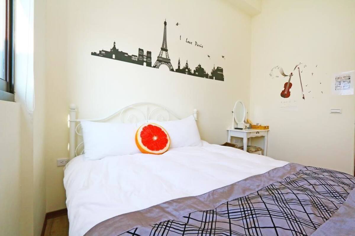 A neatly made bed with a white headboard, a single white pillow, and a red grapefruit on the pillow. There is a wall decoration of the Eiffel Tower and a violin.