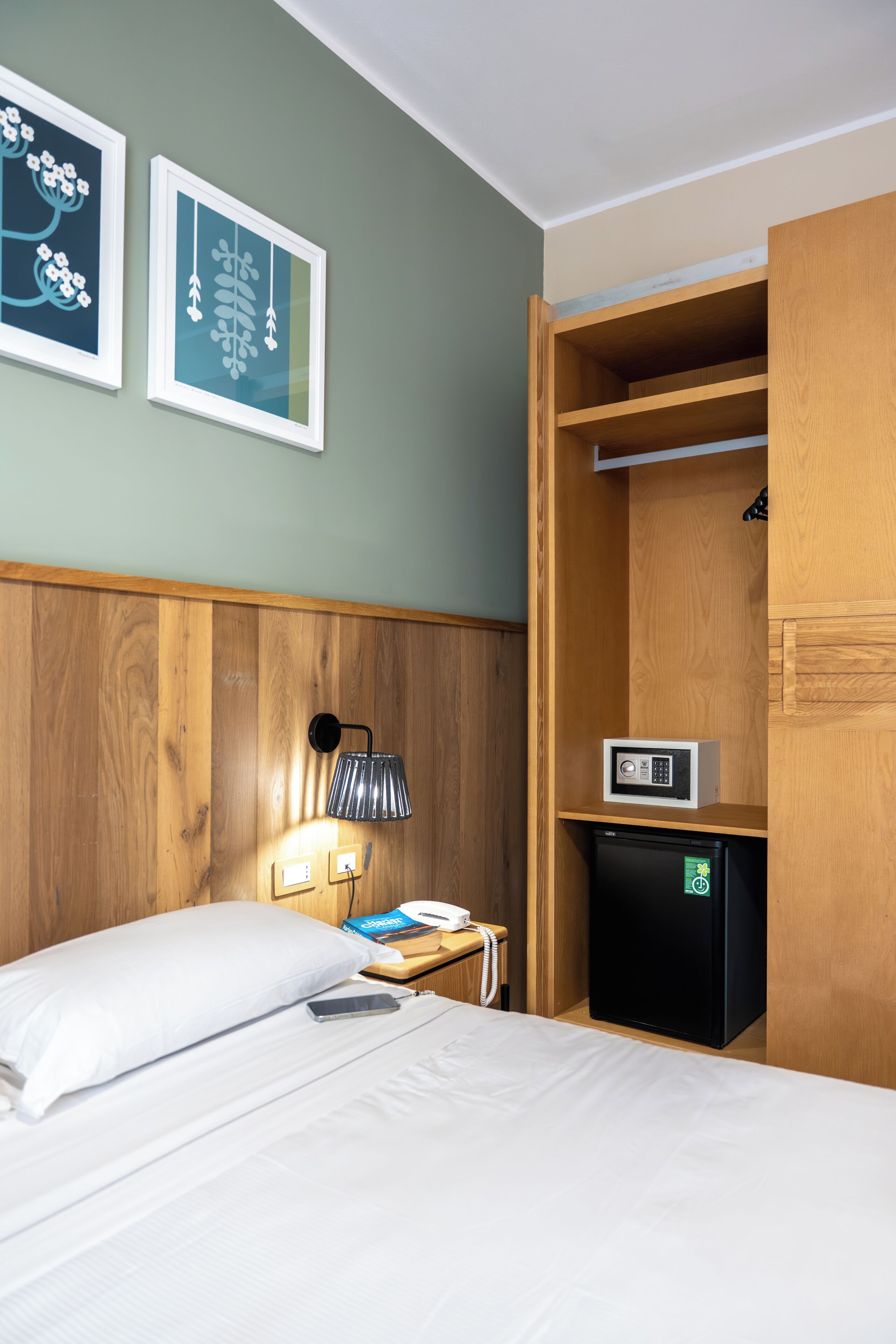 Une chambre d’hôtel avec un lit, une table de chevet avec une lampe, un téléphone, un petit réfrigérateur noir et une armoire en bois.