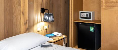 Une chambre d’hôtel avec un lit, une table de chevet avec une lampe, un téléphone, un petit réfrigérateur noir et une armoire en bois.