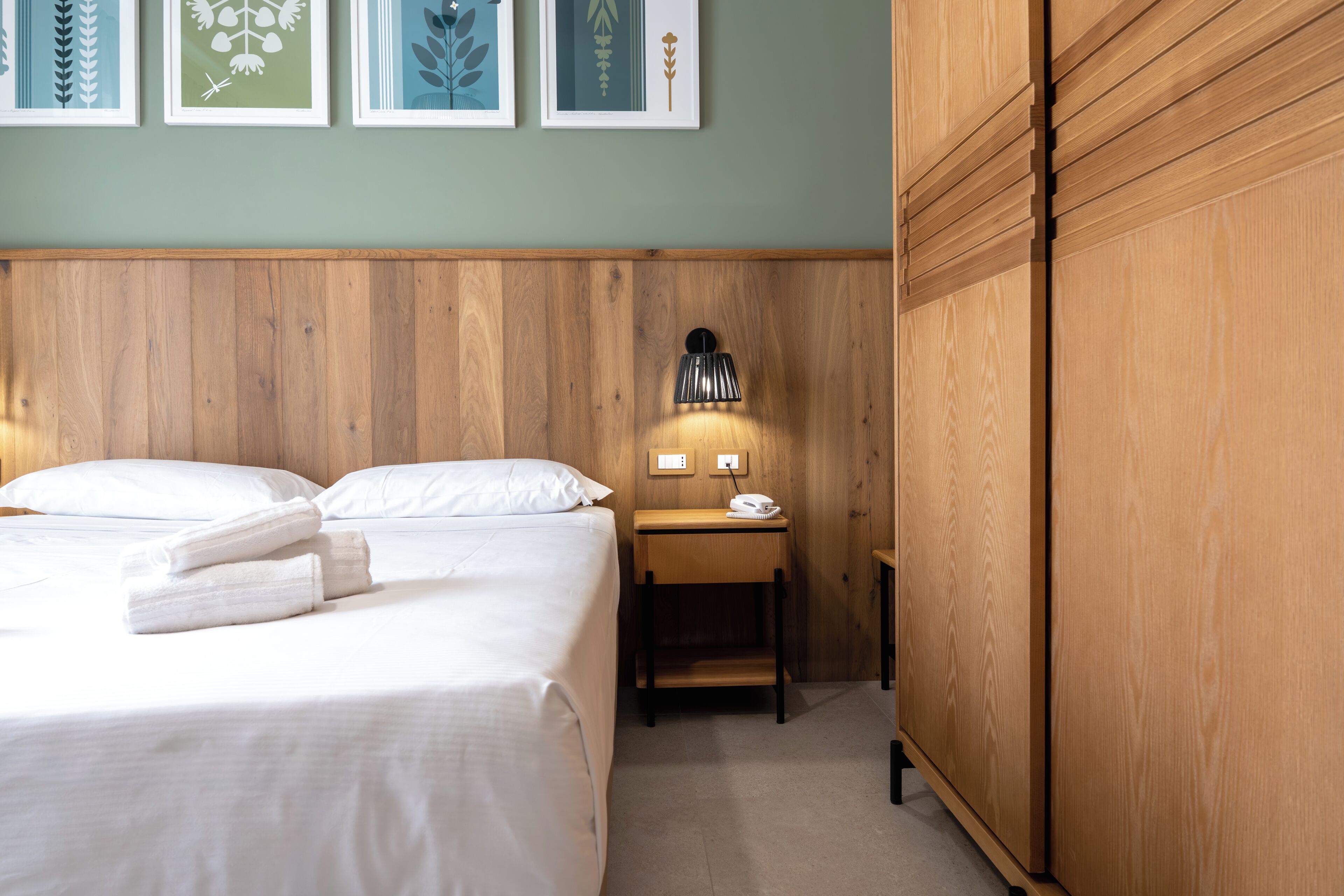 A hotel room with a wooden headboard, a bed with white linens, a bedside table with a lamp, and framed artwork on the wall.