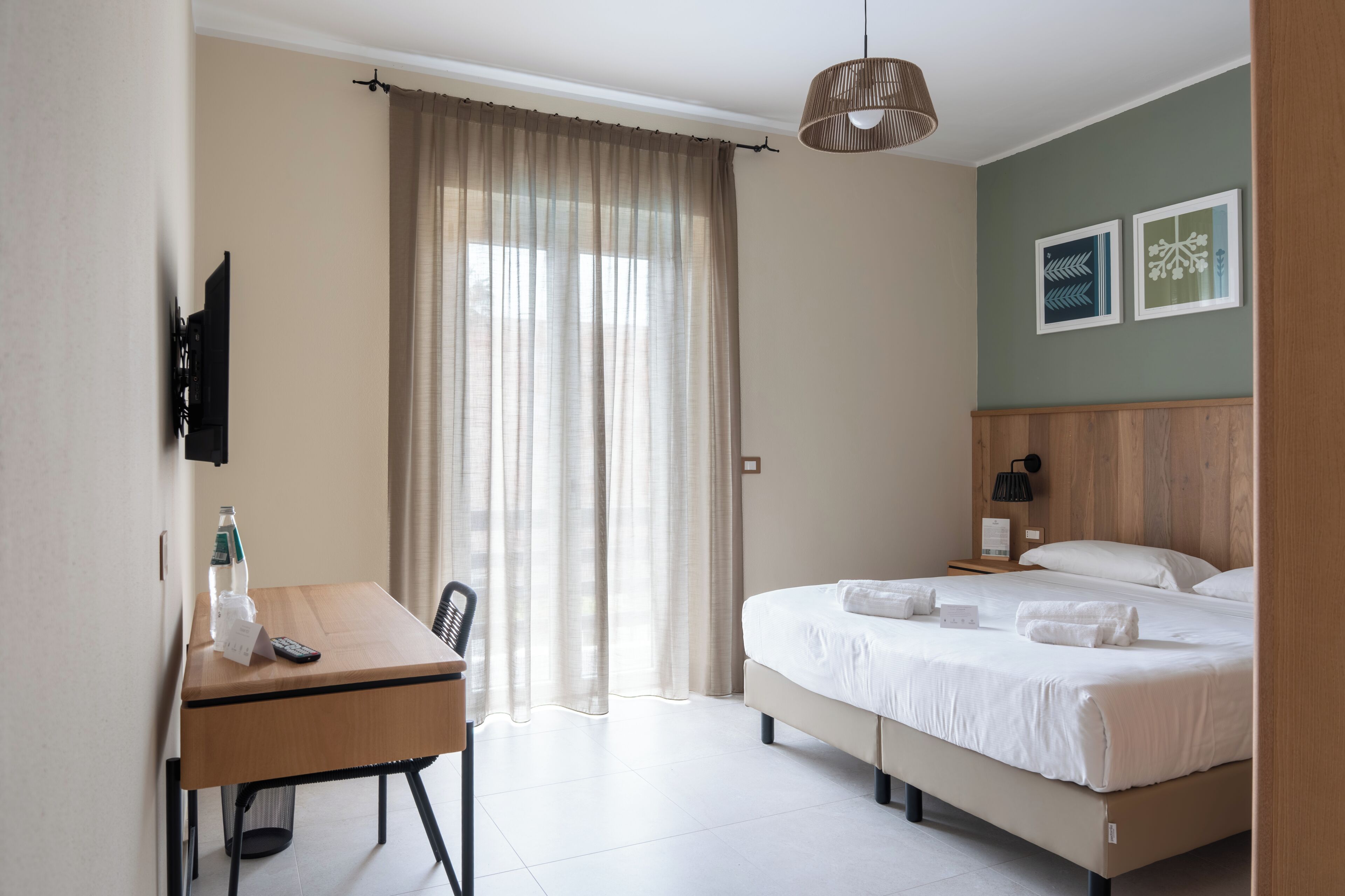 Una habitación de hotel con cama, escritorio con silla, televisor, botella de agua y cortina.