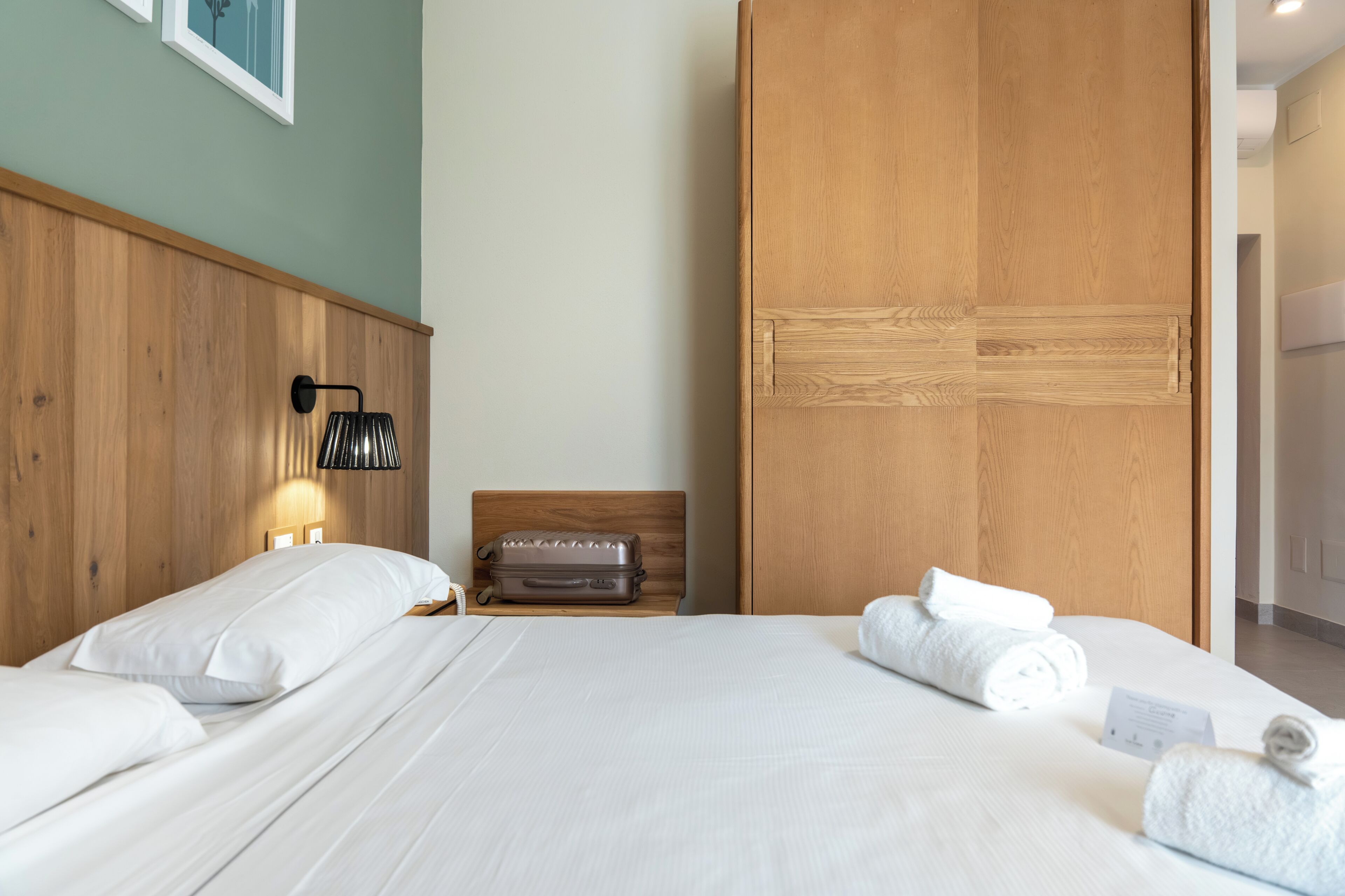 Een hotelkamer met een houten hoofdeinde, een bed met witte lakens, een koffer en opgerolde handdoeken op het bed.