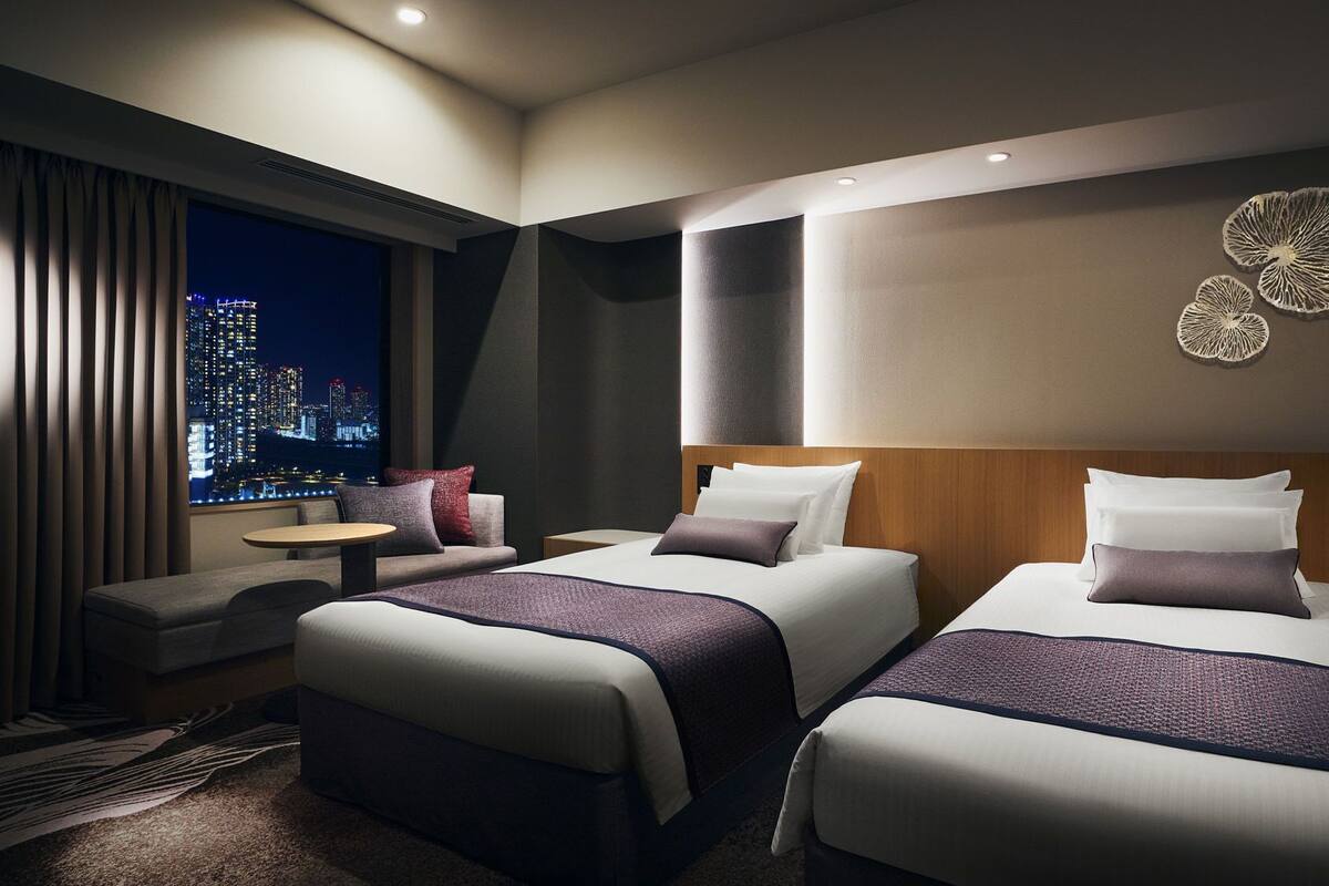 Een hotelkamer met twee bedden, een bank, een kleine tafel en uitzicht op de stad.