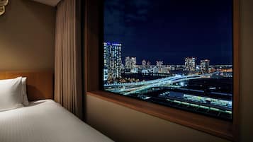 A hotel room with a large window offering a nighttime cityscape view, a neatly made bed, and a wooden headboard.
