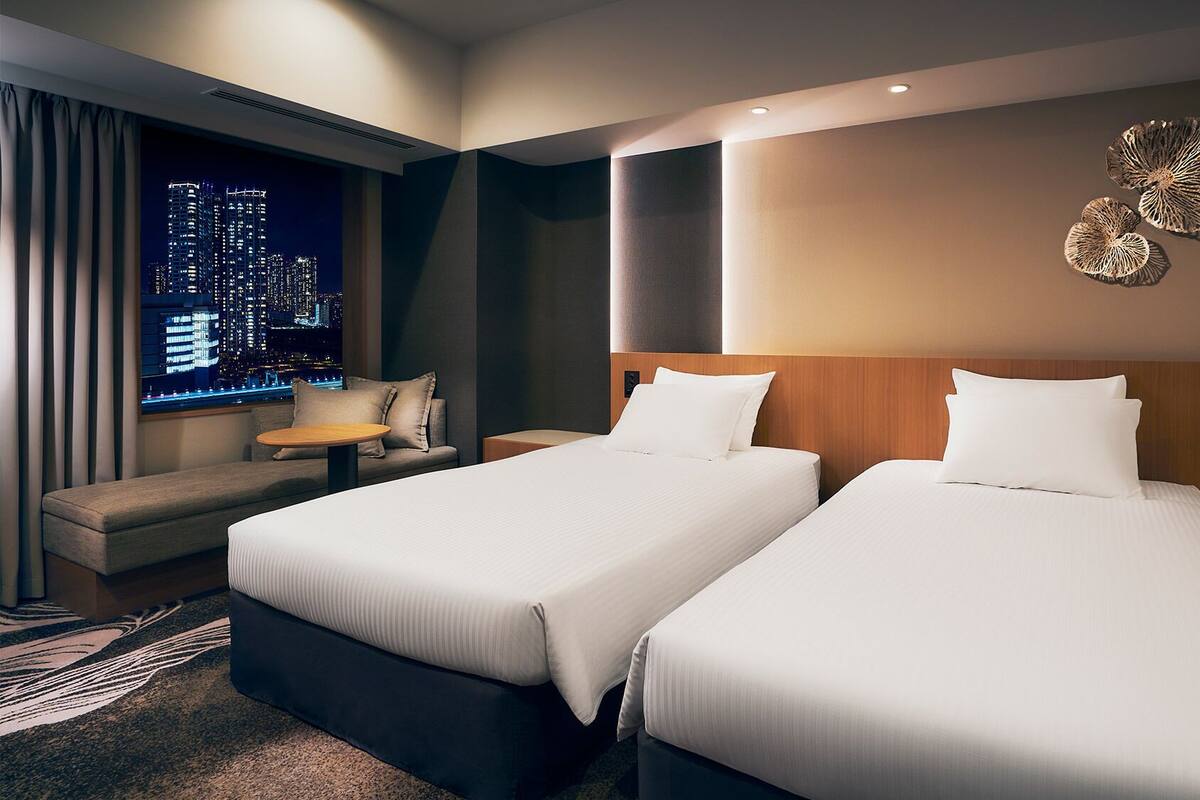 Een hotelkamer met twee bedden, een sofa, een kleine tafel en uitzicht op de stad.