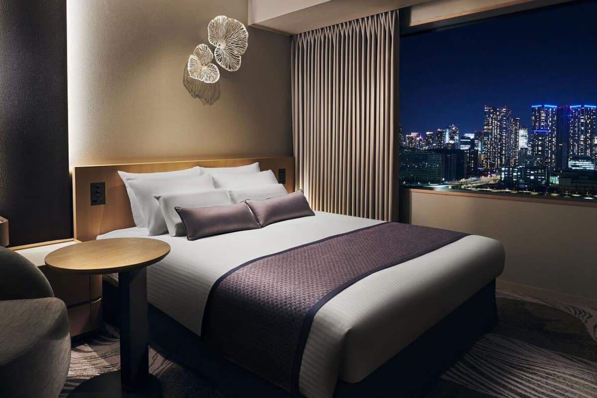 Ein Hotelzimmer mit einem großen Bett, einem runden Tisch, einem Stuhl, Blick auf die Stadt und einer Wanddekoration.