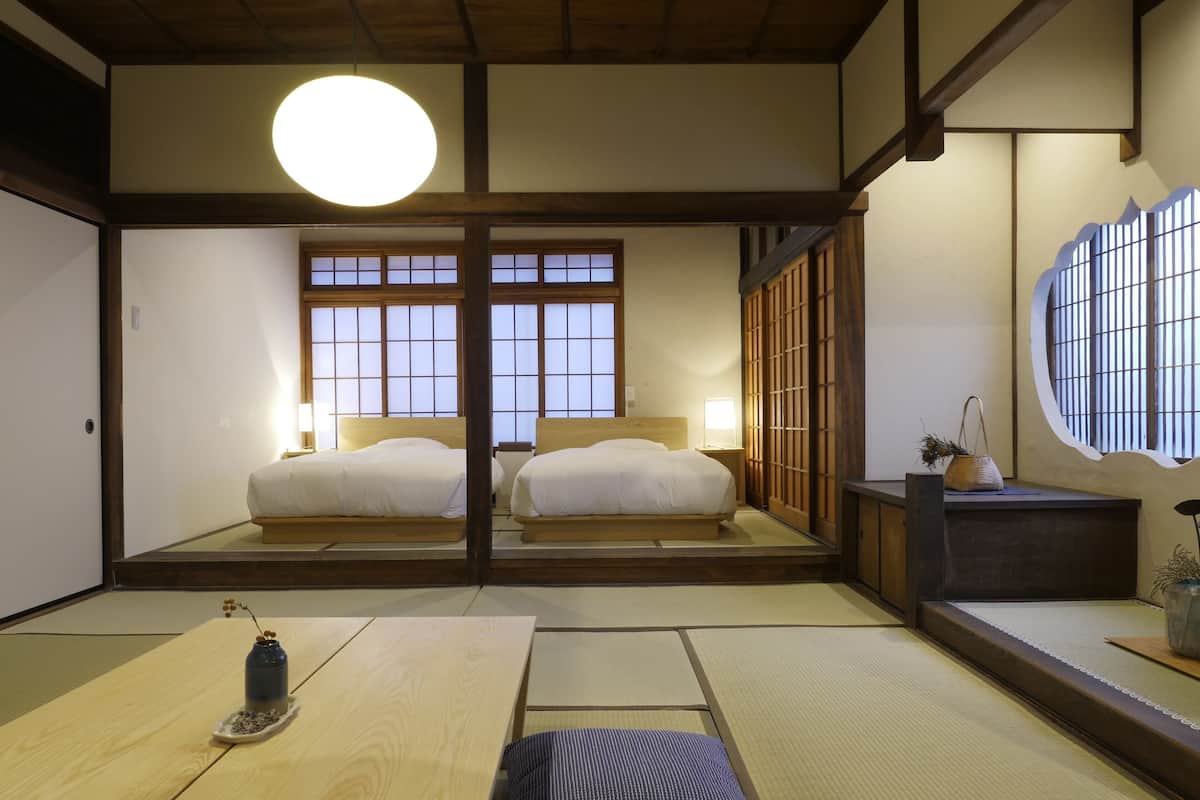 Una habitación tradicional japonesa con suelo de tatami, dos camas, una lámpara redonda grande en el techo y puertas correderas.