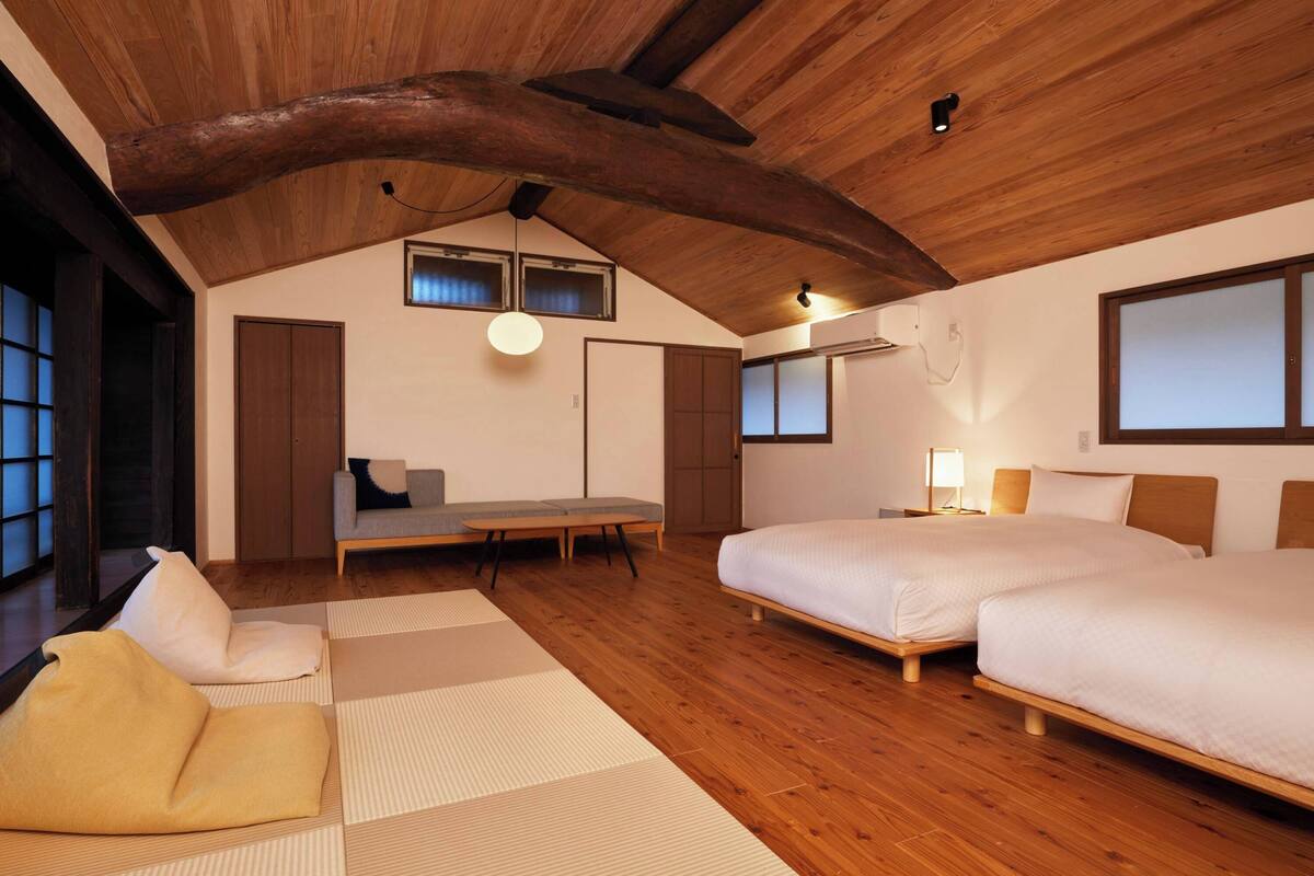 Una habitación con techo inclinado, vigas de madera, un sofá, una cama, una mesa y un ventanal.