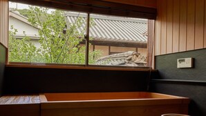 A traditional Japanese room with tatami flooring, a wooden washbasin, a window with a view of a traditional roof, and a small wooden stool.