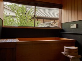 Una habitación japonesa tradicional con suelo de tatami, un lavamanos de madera, una ventana con vista a un tejado tradicional y un pequeño taburete de madera.