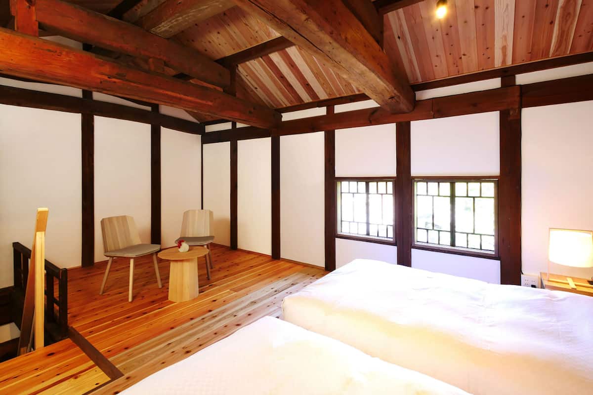 Una habitación tradicional japonesa con suelo de tatami, techo de madera bajo, una cama, un escritorio con una silla y una ventana con barrotes.