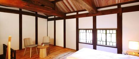 Una habitación tradicional japonesa con suelo de tatami, techo de madera bajo, una cama, un escritorio con una silla y una ventana con barrotes.