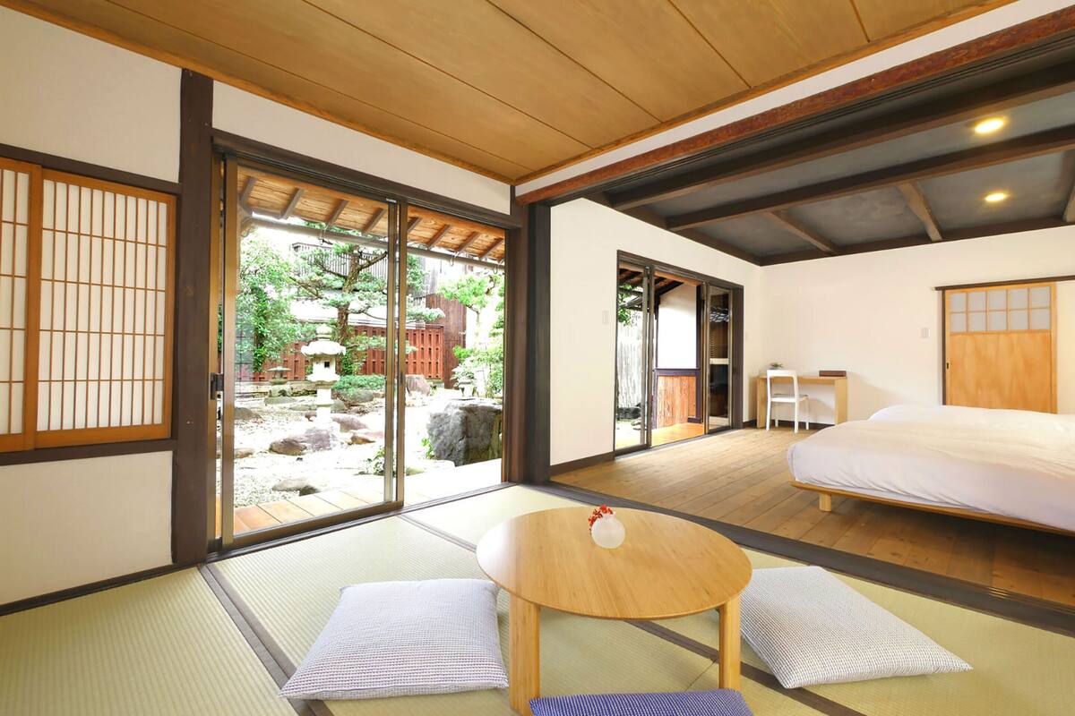 Una habitación japonesa tradicional con suelo de tatami, una cama, una mesa y puertas correderas que dan a un jardín.