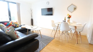 A modern living room with a black leather sofa, a dining table with white chairs, a flat-screen TV mounted on the wall, and a large window with curtains. - O2 Mailbox Central Premier Apartment (Birmingham)