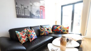 A modern living room with a black leather sofa, colorful geometric pillows, a white coffee table, and a framed cityscape painting. - O2 Mailbox Central Premier Apartment (Birmingham)