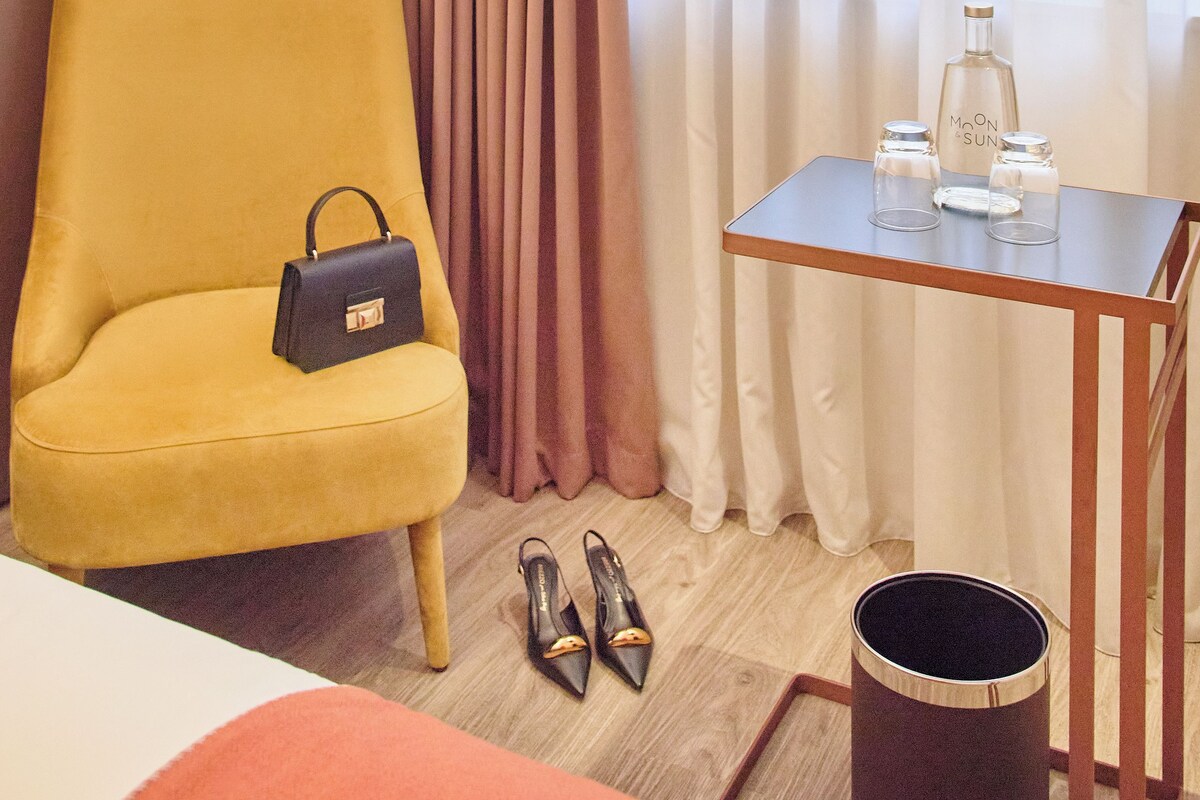 Une chambre d’hôtel avec un fauteuil jaune, une petite table sur laquelle se trouvent une bouteille et des verres, et une paire de chaussures posées sur le sol.