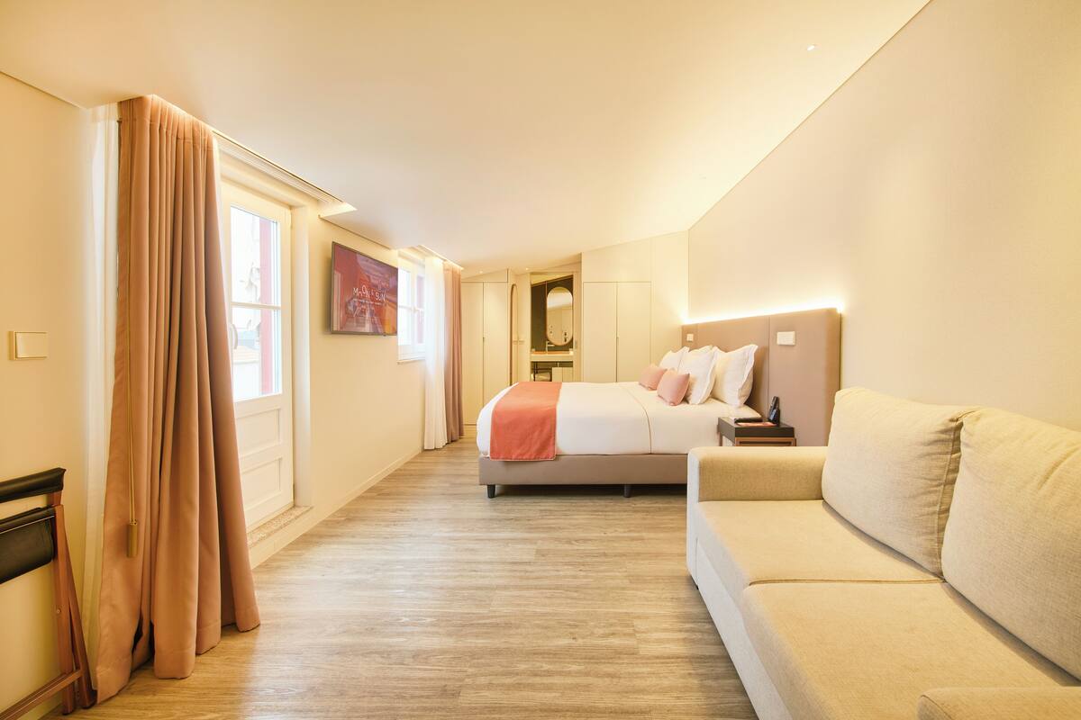 Ein Hotelzimmer mit Bett, Sofa, Holzboden, einem Fenster mit Vorhängen, einem Spiegel und einem an der Wand befestigten Fernseher.