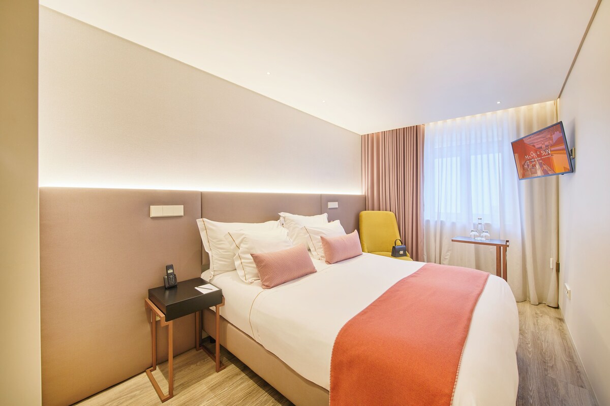 Una habitación de hotel con cama, mesita de noche, silla, televisor y cortinas.